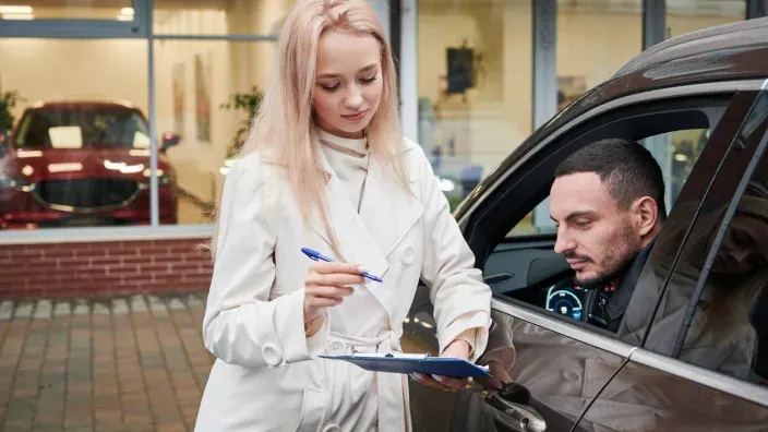 Agente comercial entregando contrato de seguro de coche o alquiler a un conductor frente a un concesionario.