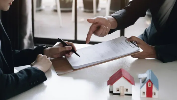 Firma de contrato de seguro de hogar o hipoteca con maquetas de casas sobre un escritorio de oficina.