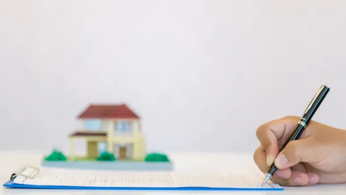 Persona firmando un contrato de compraventa o hipoteca de una vivienda, con una casa en miniatura de fondo.