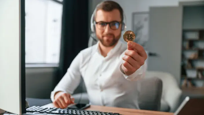 Operador de soporte técnico o broker mostrando una moneda física de Bitcoin a cámara.