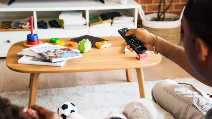 Primer plano de una mano con el mando a distancia, mientras una familia ve la tele.