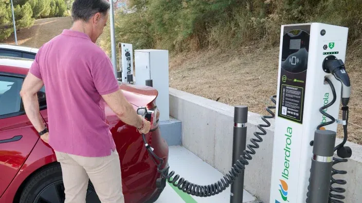 Vehículo eléctrico blanco conectado a un cargador de Iberdrola junto a una zona ajardinada.