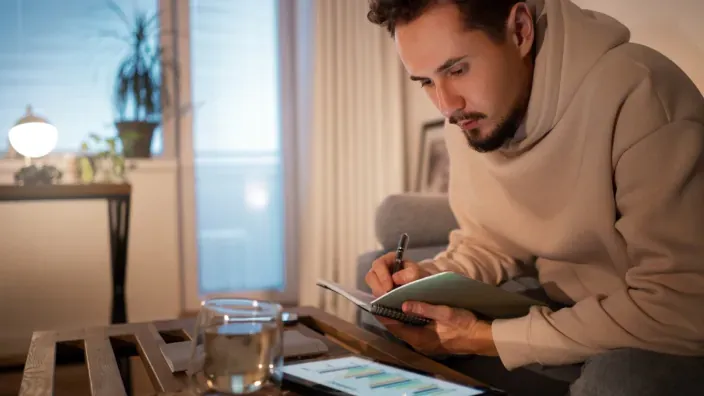 Hombre revisando sus finanzas personales en casa mientras toma notas con una tablet y gráficos en pantalla.