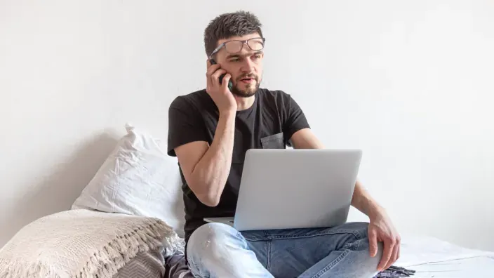 Hombre teletrabajando en casa con portátil y hablando por el móvil, representando gestiones online o soporte.