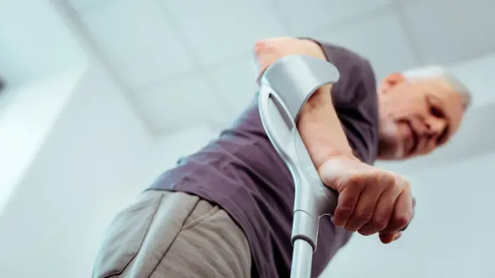 Hombre caminando con esfuerzo y apoyo de una muleta por problemas de movilidad.