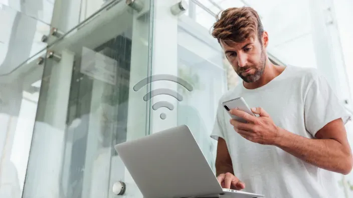 Hombre usando un portátil y su smartphone con un icono de señal Wi-Fi para representar la conectividad de internet en casa.