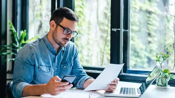Hombre con gafas revisando documentos y su smartphone, gestionando sus finanzas o trabajo desde su escritorio con un portátil.