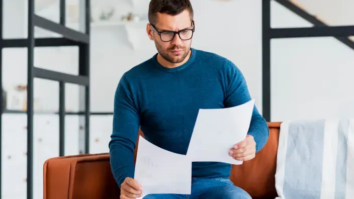 Hombre con gafas revisando facturas o documentos financieros con preocupación en el salón de su casa.