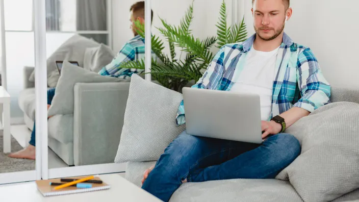 Hombre joven trabajando en el sofá de su casa con ordenador portátil y auriculares inalámbricos.