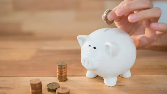 Mano introduciendo una moneda en una hucha con forma de cerdito blanco junto a pilas de monedas sobre una mesa.