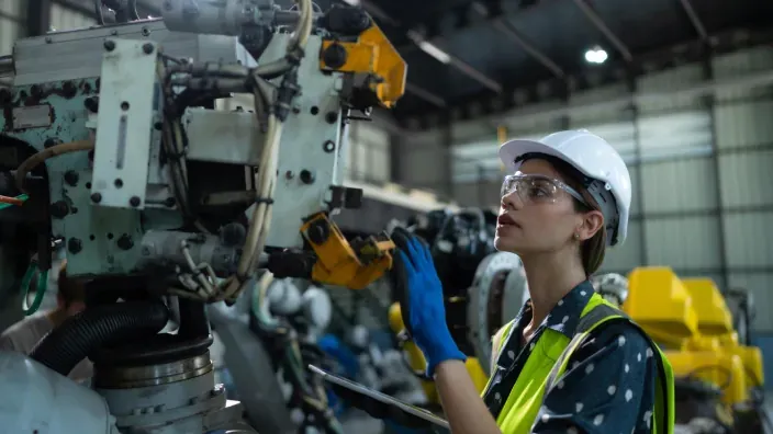 Ingeniera industrial con casco blanco y gafas de protección inspeccionando maquinaria en fábrica.