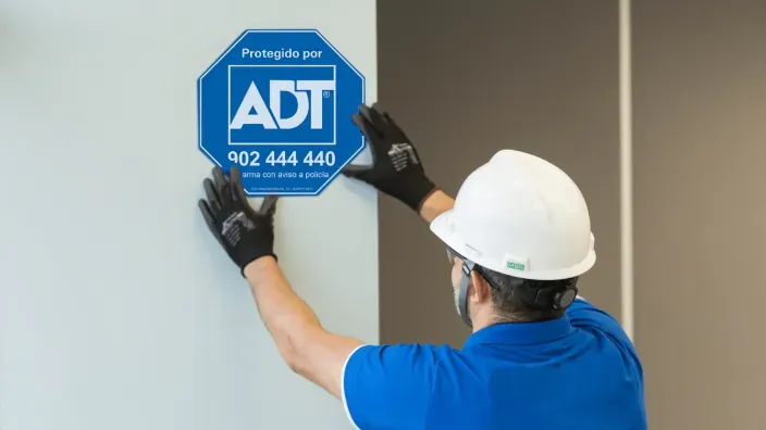 Técnico de seguridad instalando el cartel de protegido por ADT en una fachada para la vigilancia de hogares y negocios.