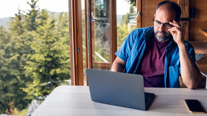 Hombre trabajando con portátil en casa rural con grandes ventanales con vistas al bosque.