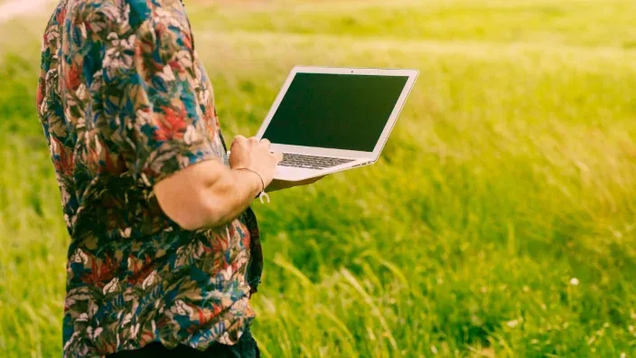 Hombre usando un ordenador portátil en el campo para representar el teletrabajo y la conectividad en zonas rurales.