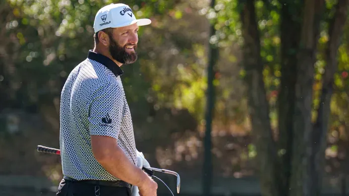 El golfista español Jon Rahm, en el campo con gorra blanca y polo estampado sosteniendo un palo de golf.