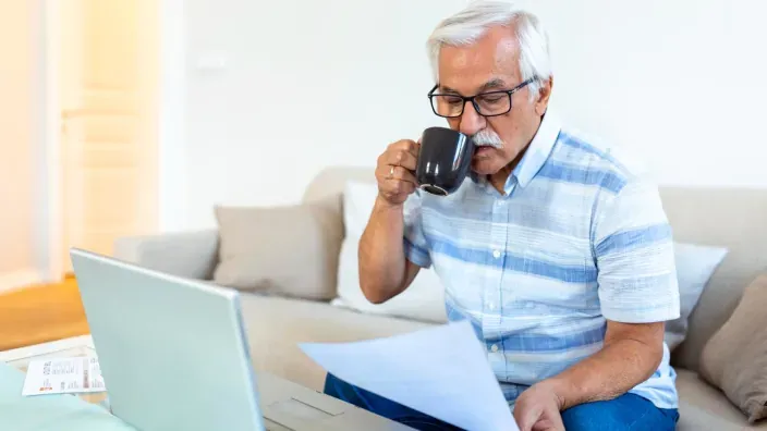 Hombre jubilado revisando documentos y facturas en su portátil mientras toma café en el sofá de su hogar.