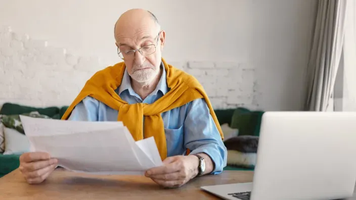 Hombre mayor leyendo documentos financieros frente a un ordenador portátil en casa para planificar su jubilación.