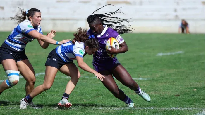 Jugadora de rugby femenino corre con el balón mientras dos rivales intentan placarla. Partido de la División de Honor.