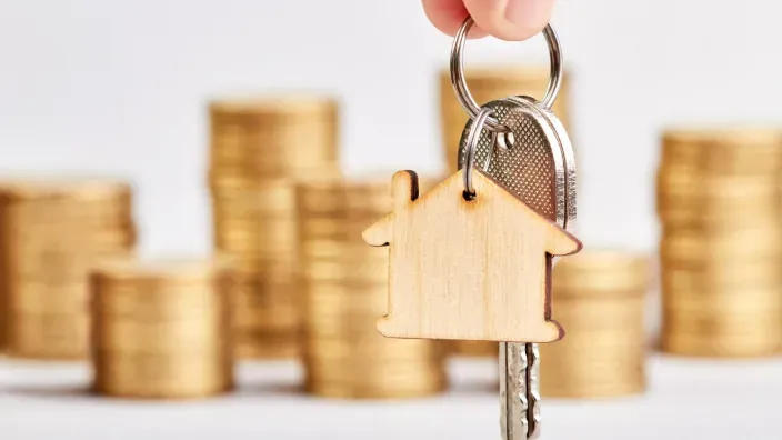 Llaves con llavero en forma de casa frente a monedas apiladas simbolizando ahorro para vivienda.