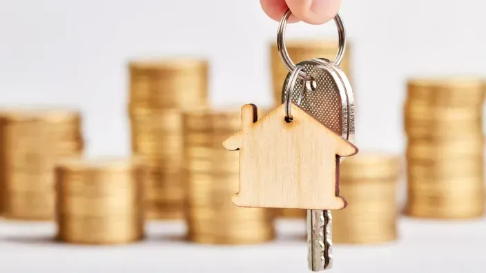 Llaves de casa con un llavero en forma de vivienda delante de varias pilas de monedas