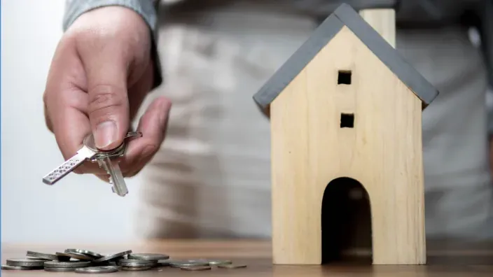 Mano sosteniendo unas llaves junto a una casita de madera y monedas sobre la mesa.