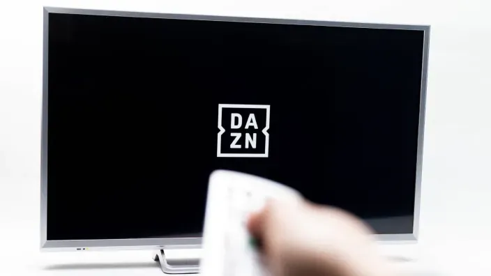 Mano con un mando a distancia apuntando a un televisor con el logo de la plataforma de streaming DAZN en la pantalla.