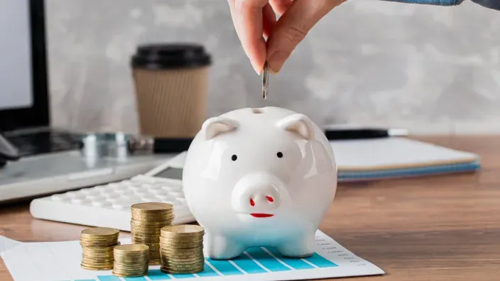 Mano introduciendo una moneda en una hucha con forma de cerdito sobre una mesa.