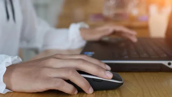 Primer plano de manos de mujer haciendo una consulta con ratón inalámbrico y portátil en escritorio.