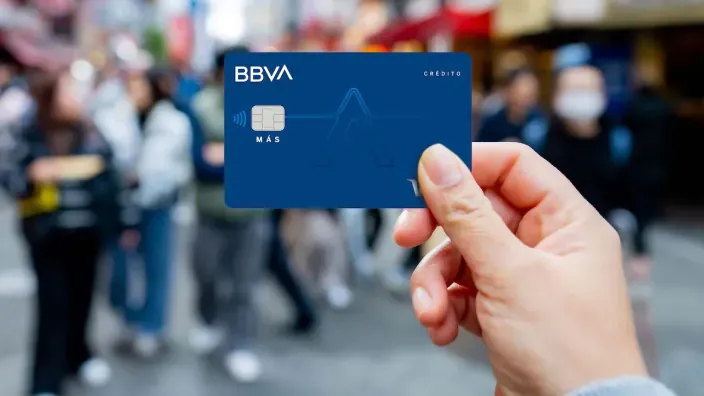 Mano sosteniendo una tarjeta azul de BBVA en una calle con personas desenfocadas al fondo