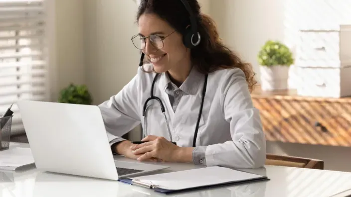 Médica con auriculares y fonendoscopio trabajando ante un ordenador portátil