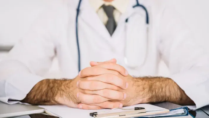 Primer plano de un médico con bata blanca y estetoscopio, con las manos entrelazadas sobre la mesa.