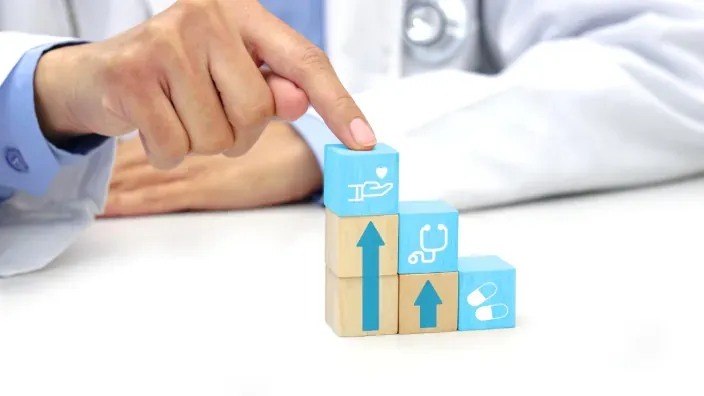 Mano de médico señalando bloques de madera con iconos de salud y flecha de crecimiento ascendente.