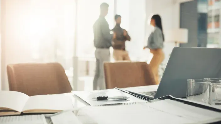 Planificación en oficina: documentos y portátil en primer plano con plantilla de trabajadores hablando al fondo.