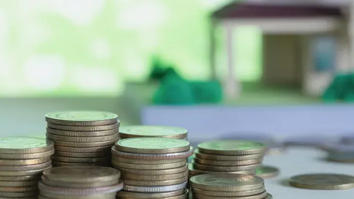 Montones de monedas apiladas con una maqueta de casa desenfocada al fondo.