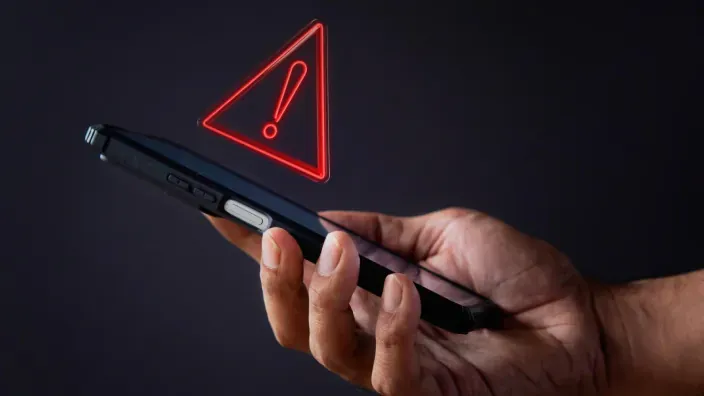 Smartphone con un icono de advertencia rojo sobre la mano de un usuario para representar alertas de ciberseguridad.