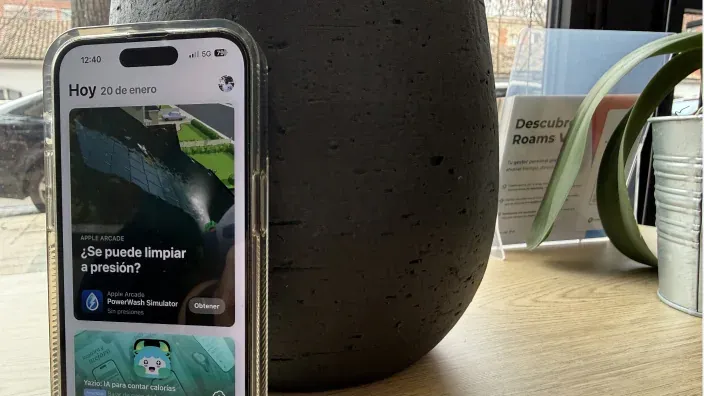 Teléfono móvil apoyado sobre una mesa mostrando una pantalla con la app store abierta, junto a una maceta y material informativo en una oficina.