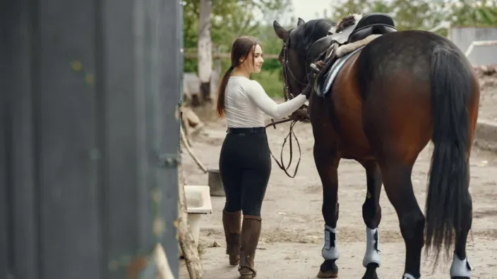 Mujer caminando junto a un caballo ensillado en el exterior de una cuadra.