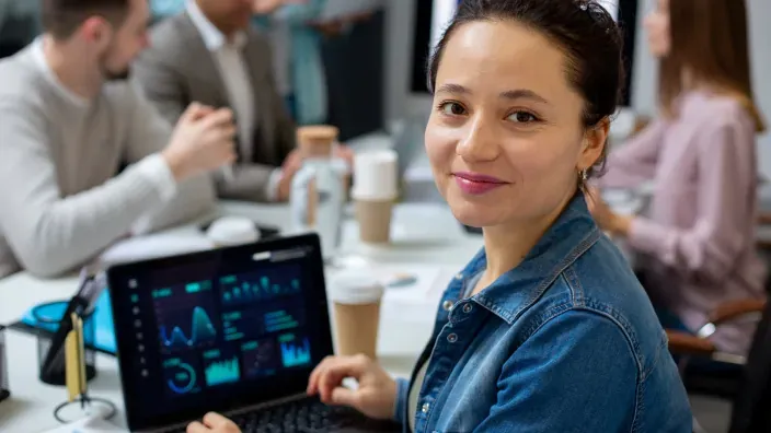 Mujer analista de datos sonriendo en la oficina con un portátil que muestra gráficos y métricas de negocio.