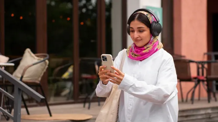Mujer joven musulmana con hijab rosa y auriculares negros usando smartphone caminando por la calle.