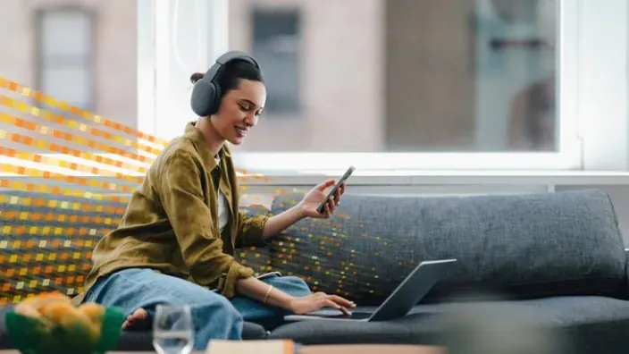 Mujer con auriculares usando un portátil y un móvil en el sofá para representar la conectividad de fibra óptica en casa.