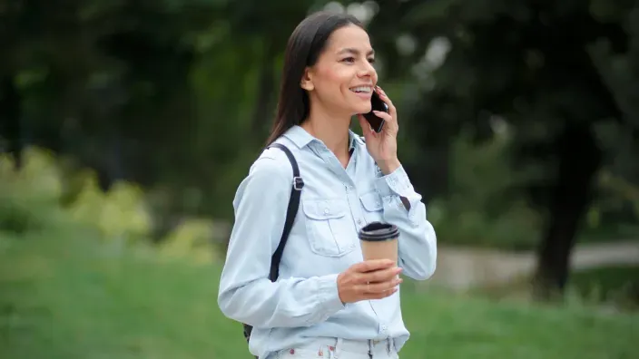 Mujer hablando por teléfono móvil y sosteniendo un café para llevar mientras pasea por un parque urbano.