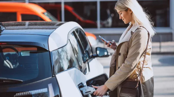 Mujer cargando un coche eléctrico en la vía pública mientras consulta el móvil para controlar la recarga.