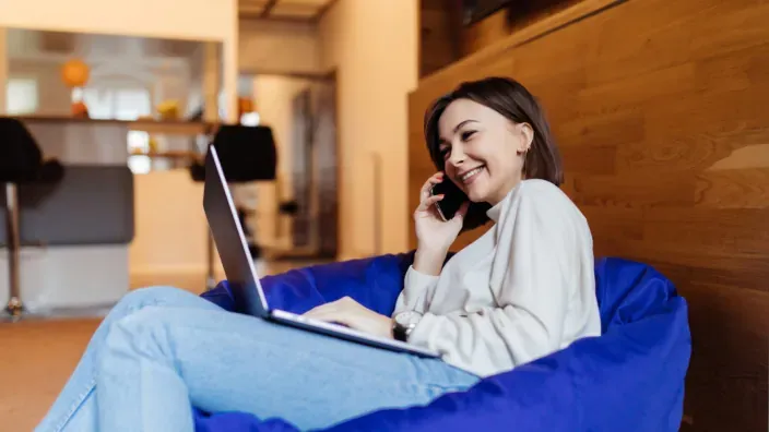 Joven teletrabajando en un puff azul con un ordenador portátil y hablando por smartphone en un ambiente relajado.
