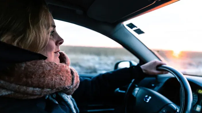 Mujer conduciendo un coche al atardecer, luciendo bufanda de invierno, imagen que ilustra la preparación y seguridad en la conducción ante bajas temperaturas.