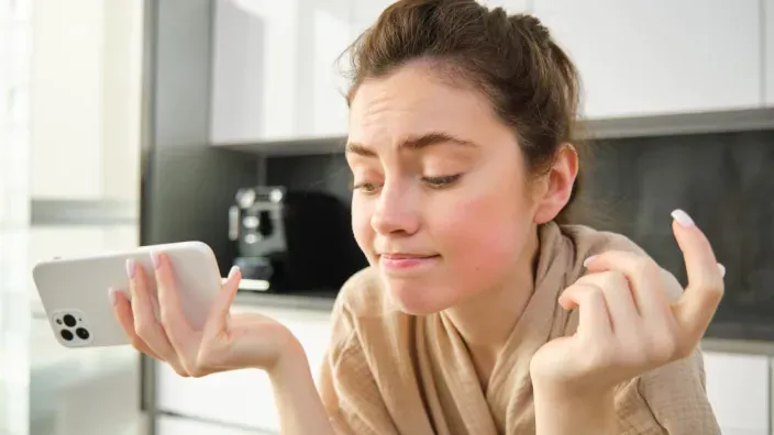 Mujer joven con gesto de duda o frustración mirando su móvil. Problemas con el servicio, la cobertura o la velocidad de internet.