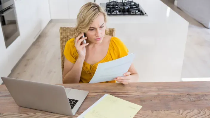 Mujer con gesto de enfado hablando por teléfono mientras revisa una factura o contrato, con su ordenador portátil al lado.