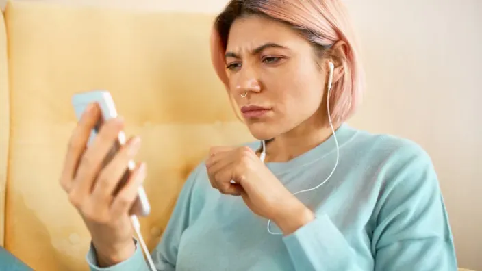 Joven con auriculares mirando su smartphone con gesto de duda o preocupación por una consulta online.