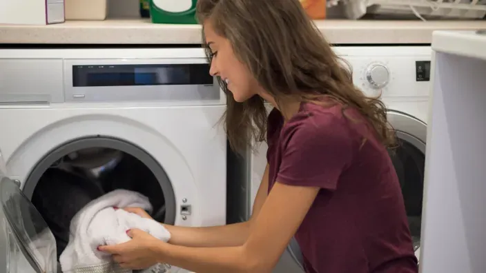 Mujer sonriendo mientras introduce ropa en una lavadora en un entorno doméstico.