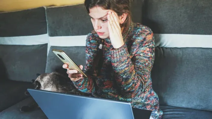 Chica preocupada mirando el móvil con mano en la frente frente al ordenador portátil.
