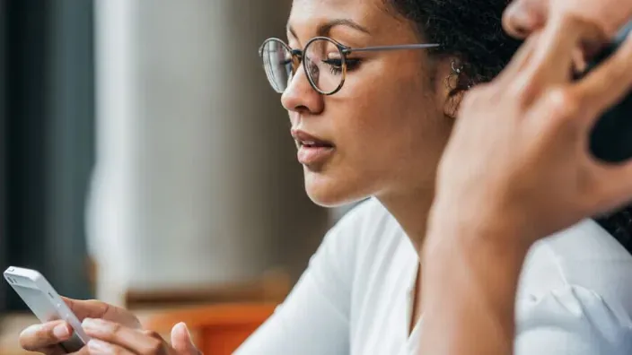 Mujer con gafas consultando su smartphone en la oficina junto a otra persona hablando por teléfono al fondo.